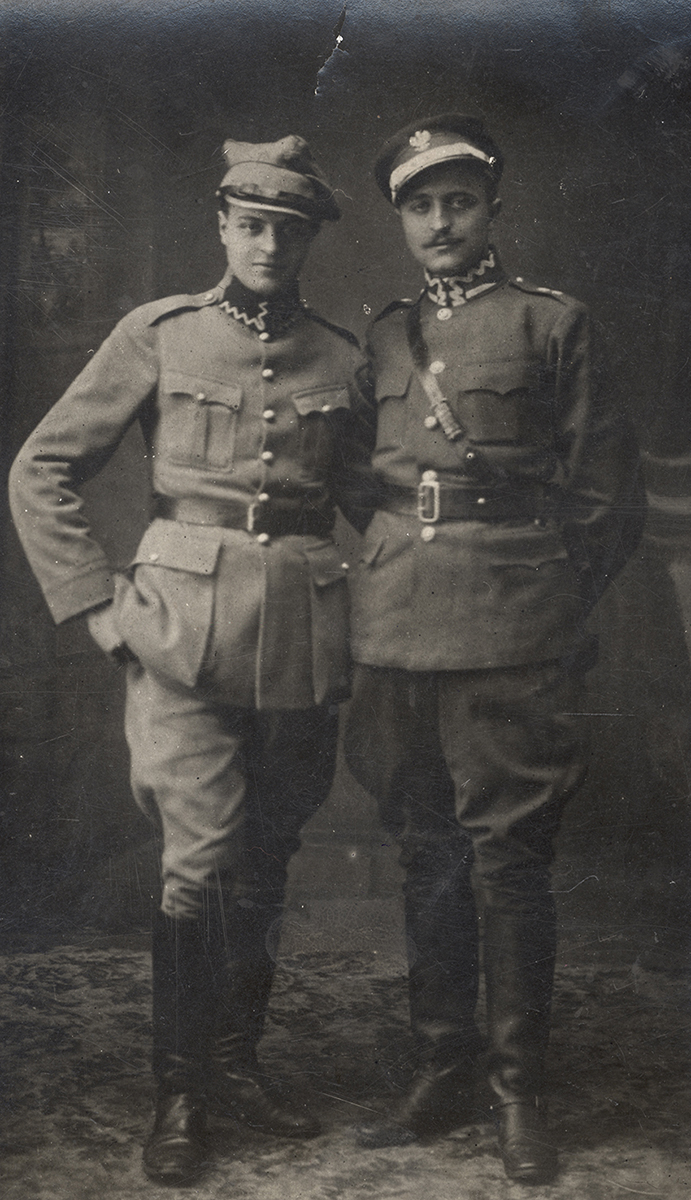 Fotografia archiwalna czarno-biała, zrobiona około 1920 roku, przedstawia dwóch młodych mężczyzn, ujętych w całej postaci, ubranych w mundury wojska polskiego. Pozują na ciemnym tle fotograficznym.