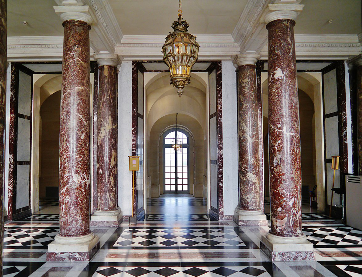 Fotografia kolorowa, współczesna, przedstawiająca wnętrze Westybulu Marmurowego w pałacu w Wersalu. Jasne pomieszczenie z czterema marmurowymi kolumnami w odcieniach brązu i wysokimi przejściami. W głębi, pośrodku widoczne zakończone półkoliście okno podzielone listwami. Przy suficie złocona latarnia w kształcie odwróconej gruszki z przeszkleniem w postaci kwadratowych szybek.