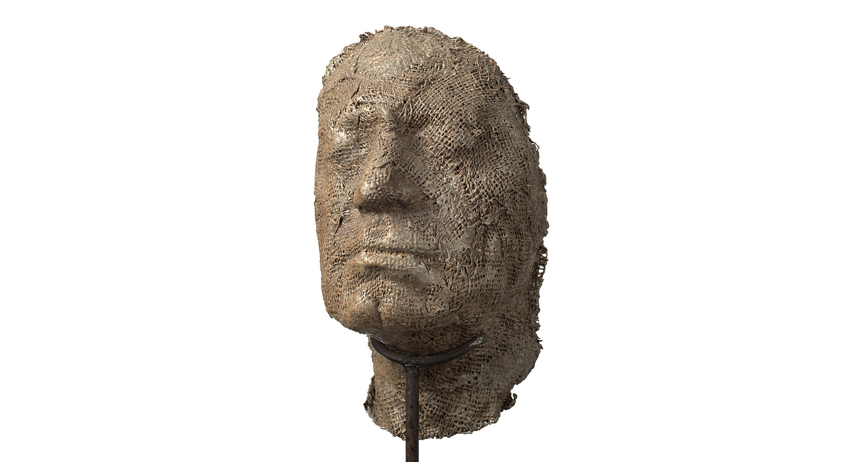 Magdalena Abakanowicz, Portret anonimowy, © Fundacja Marty Magdaleny Abakanowicz Kosmowskiej i Jana Kosmowskiego Magdalena Abakanowicz
urodzona w 1930 roku w Falentach – zmarła w 2017 roku w Warszawie
Portret anonimowy
powstały w 2000 roku
Żywica metal (stelaż)
wymiary: 103 x 22 x 23
Starak Collection
Praca ukazuje silnie uproszczoną, niemal ascetyczną głowę – rzeźbę wykonaną z żywicy o powierzchni przypominającej spękaną, wyschniętą ziemię. Materiał ten nadaje formie porowatą, kruchą strukturę, która zdaje się być świadectwem upływu czasu, przemijania i organicznej ulotności.
Głowa jest zamocowana na długim, cienkim metalowym pręcie, który osadzony jest w minimalistycznej, kwadratowej podstawie. Ten kontrast między miękką, niemal organiczną materią a zimnym, surowym metalem podkreśla napięcie między cielesnością a abstrakcją, między ludzkim doświadczeniem a mechaniczną formą nośną.
Twarz – pozbawiona większości szyi, jakby wyjęta z ciała i zawieszona w przestrzeni – emanuje głęboką emocją. Zamknięte lub przymknięte oczy otoczone są wyraźnymi bruzdami, które mówią o cierpieniu, melancholii lub intensywnej zadumie. Usta lekko opuszczone podkreślają powagę, a może nawet ból, tworząc wyraz pełen dramatyzmu i skupienia.
Rzeźba zdaje się nie tylko przedstawiać postać, ale raczej wprowadzać widza w przestrzeń emocjonalną, gdzie surowość materiału i minimalizm formy stają się nośnikiem uniwersalnej ludzkiej kondycji.