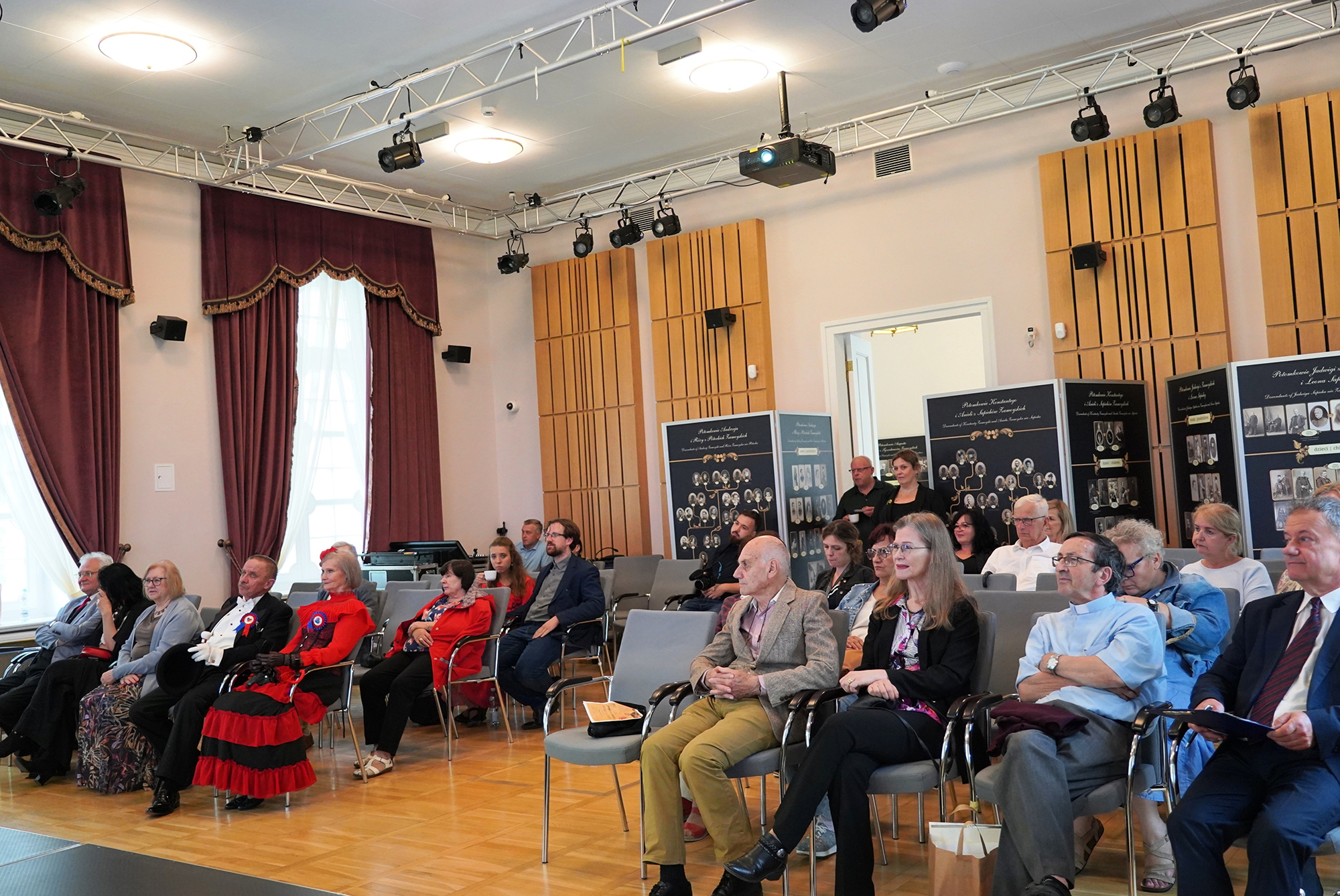 Fot. 2 Miejsce akcji to elegancka sala konferencyjno-wystawowa z wysokim sufitem i dębową podłogą. W pomieszczeniu zgromadzone jest około 30 osób, które siedzą w szarych krzesłach i zwrócone są w kierunku, z którego wykonano zdjęcie – prawdopodobnie scena lub mównica.
Po lewej stronie sali wiszą ciemnoczerwone, ciężkie zasłony z dekoracyjnymi lambrekinami, a z sufitu zwisają światła sceniczne i projektory zamocowane na metalowych belkach.
Większość obecnych to osoby dorosłe i starsze, elegancko ubrane – w garnitury, sukienki, marynarki. W oczy rzuca się para ubrana w stroje przypominające historyczne kostiumy: mężczyzna w czarnym fraku z białymi rękawiczkami i kobieta w czerwono-czarnej sukni stylizowanej na XIX-wieczną.
W tle, po prawej stronie sali, stoją plansze wystawowe przedstawiające stare fotografie, portrety i podpisy – prawdopodobnie fragment wystawy historycznej lub genealogicznej. Nad nimi widnieją napisy w języku polskim. Wśród zgromadzonych widać skupienie, zainteresowanie i ciszę – panuje podniosła atmosfera, typowa dla uroczystych wydarzeń kulturalnych, naukowych lub rocznicowych.