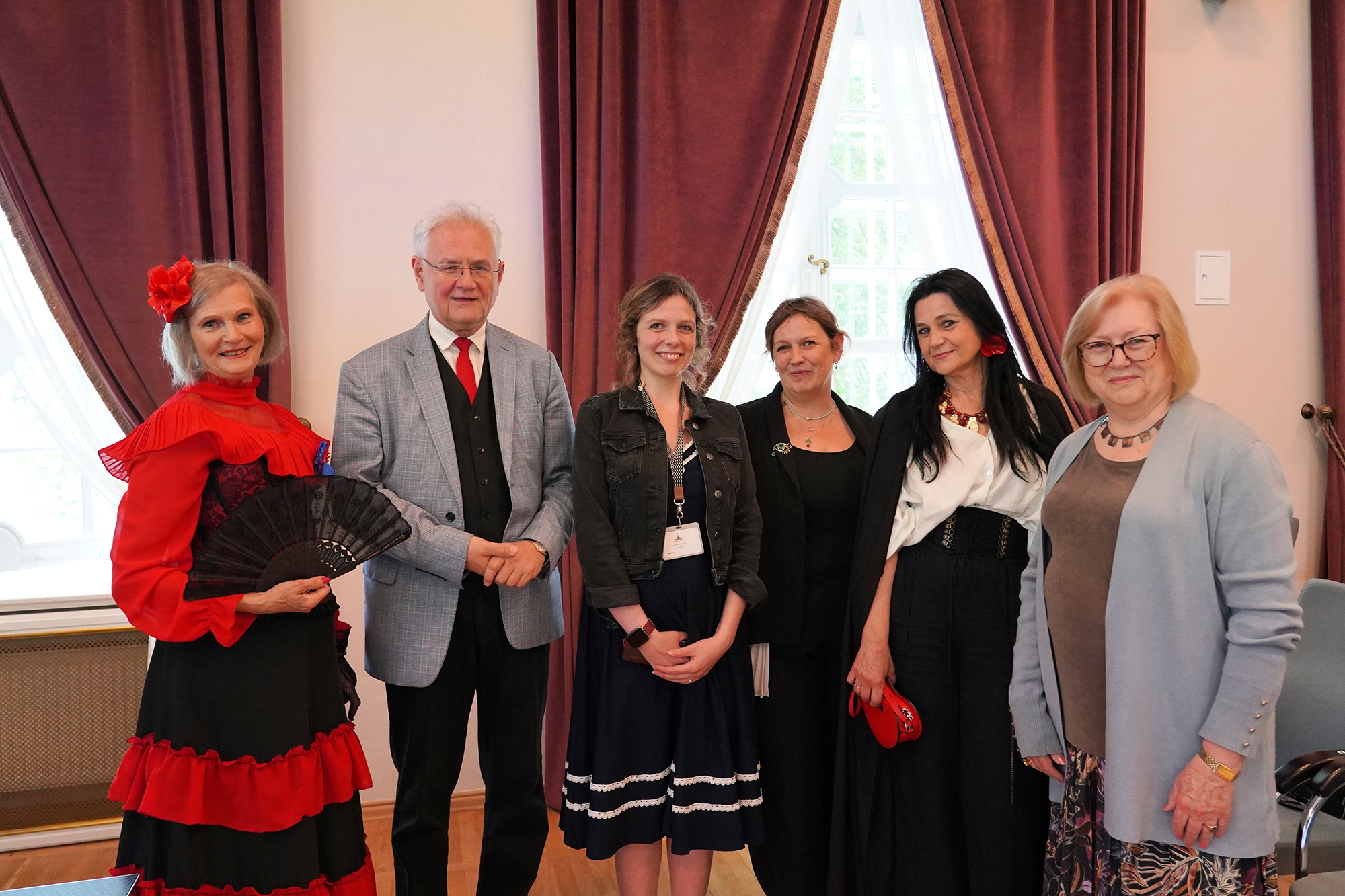 Fot. 3 Na fotografii widzimy siedem osób stojących w rzędzie i pozujących do wspólnego zdjęcia w eleganckim wnętrzu, najprawdopodobniej podczas wydarzenia kulturalnego lub konferencji. W tle znajdują się ciemnoczerwone zasłony i wysokie okna, przez które wpada naturalne światło. Pomieszczenie ma klasyczny wystrój.
Od lewej strony:
Kobieta w efektownym stroju stylizowanym na hiszpański lub XIX-wieczny – ma na sobie czarno-czerwoną suknię z falbanami, duży czerwony kwiat we włosach oraz trzyma czarny wachlarz. Uśmiechnięta.
Mężczyzna w jasnej marynarce w kratkę, ciemnej kamizelce i czerwonym krawacie – ma siwe włosy i okulary, również uśmiecha się do obiektywu.
Młoda kobieta w granatowej sukience i czarnej kurtce dżinsowej, z identyfikatorem zawieszonym na szyi. Ma rozpuszczone, kręcone włosy i delikatny uśmiech.
Kobieta w czarnej eleganckiej sukni z broszką, krótkie włosy, lekki uśmiech.
Kobieta w białej bluzce z dużym naszyjnikiem i czarnej spódnicy lub spodniach, długie ciemne włosy, trzyma małą czerwoną torebkę. Na uchu widoczna czerwona ozdoba przypominająca kwiat.
Kobieta w okularach, ubrana w szarą narzutkę i wzorzystą spódnicę – stoi po prawej stronie zdjęcia, spokojny wyraz twarzy.
Wszyscy wyglądają na zadowolonych i zaangażowanych w wydarzenie, w którym uczestniczą. Stylizacje świadczą o różnorodnym charakterze spotkania – część osób ubrana jest klasycznie, inne bardziej artystycznie lub historycznie.