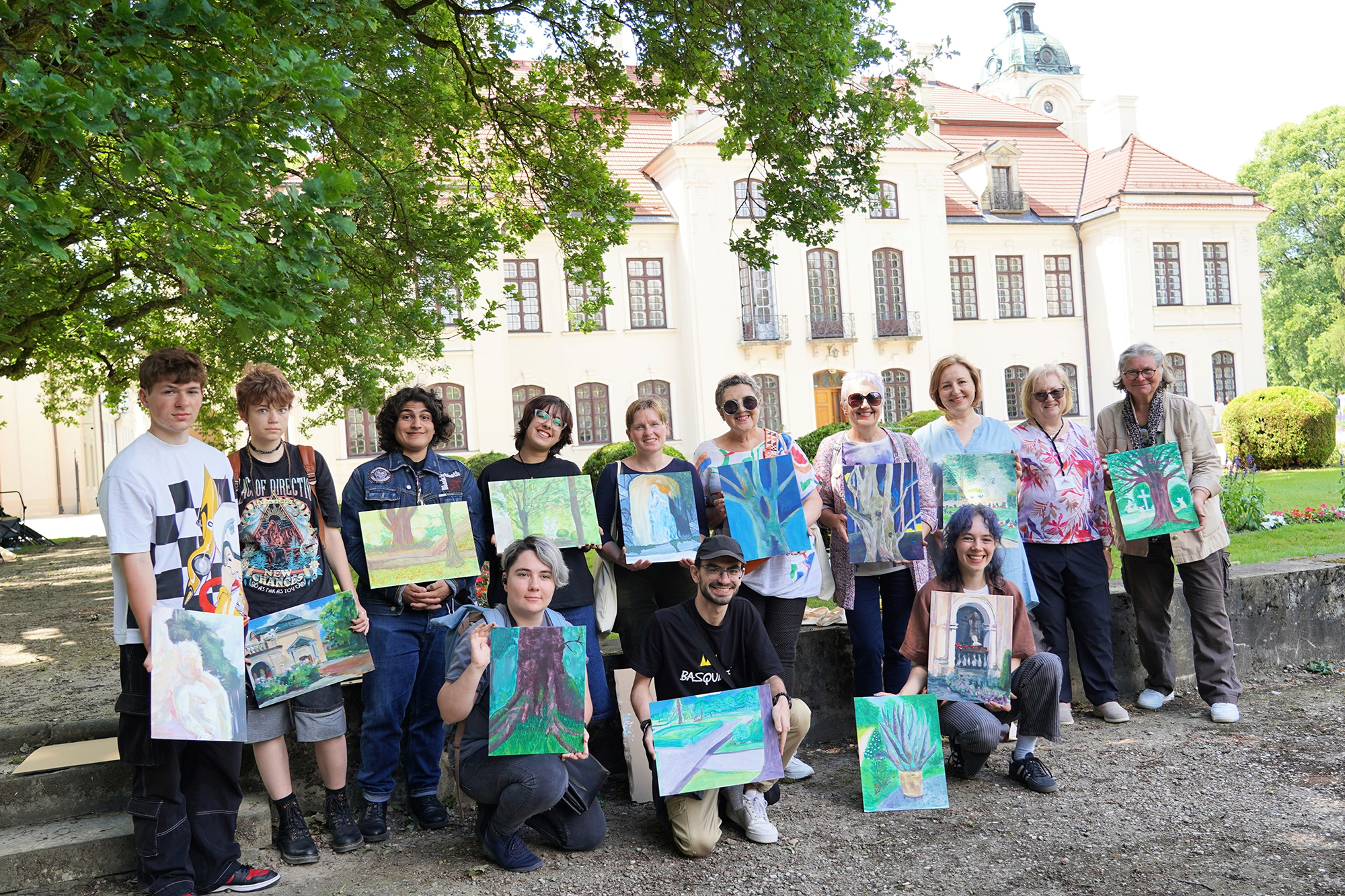 Fot. 1 Fotografia przedstawia siedem osób stojących obok siebie w jasnym, eleganckim pomieszczeniu z wysokimi oknami zasłoniętymi ciemnopurpurowymi zasłonami z ozdobnymi lambrekinami. Ściany są jasne, a przez okna wpada naturalne światło. Podłoga jest drewniana. W tle widać elementy klasycznego wystroju – grzejniki pod oknami i fragmenty krzeseł.
Od lewej strony:
Kobieta w czarno-czerwonej sukni z falbanami stylizowanej na strój hiszpański lub teatralny. Ubrana w czerwień i czerń, z dużym czerwonym kwiatem we włosach. W ręku trzyma czarny wachlarz. Uśmiecha się szeroko, patrząc w obiektyw.
Starszy mężczyzna z siwymi włosami i okularami, ubrany elegancko – ma jasnoszarą marynarkę w kratę, czarną kamizelkę, białą koszulę i czerwony krawat. Uśmiechnięty, ręce złożone przed sobą.
Młoda kobieta w granatowej sukience z białymi akcentami i czarnej kurtce dżinsowej. Na szyi zawieszony identyfikator. Ma jasnobrązowe, falujące włosy, splecione z tyłu. Uśmiecha się naturalnie.
Kobieta w czarnej, eleganckiej sukience i marynarce, z krótkimi włosami. Ma broszkę na lewej piersi i delikatny uśmiech.
Kobieta o długich, ciemnych włosach, ubrana w białą koszulę z szerokimi rękawami i czarne spodnie lub spódnicę. Ma na sobie naszyjnik i ozdobne czerwone kolczyki przypominające kwiaty. W prawej ręce trzyma małą czerwoną torebkę. Patrzy w obiektyw z lekkim uśmiechem.
Kobieta w okularach, blond włosy do ramion, ubrana w brązową bluzkę i jasnoszarą narzutkę. Ma wzorzystą spódnicę i naszyjnik. Wygląda spokojnie i dostojnie.
(Osoba częściowo widoczna za grupą – nieopisana ze względu na brak wyraźnych detali.)
Wszyscy pozują do wspólnego zdjęcia, stojąc blisko siebie, zrelaksowani i uśmiechnięci. Zdjęcie oddaje przyjazną atmosferę spotkania, być może po części oficjalnego lub kulturalnego wydarzenia.