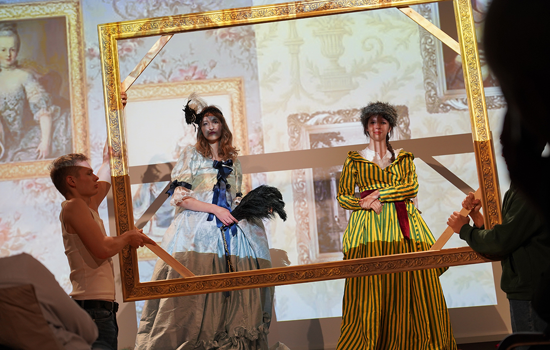 Na zdjęciu widać scenę przedstawienia inspirowanego dawnym malarstwem. Na pierwszym planie dwie młode osoby stoją wewnątrz dużej, złotej ramy obrazu, którą po bokach trzymają inni uczestnicy spektaklu.
Postać po lewej stronie ma na sobie błękitną, długą suknię z bufami i ciemnoniebieskimi wstążkami, na głowie czarną ozdobę z piórami. W dłoniach trzyma czarne pióro – wachlarz. Obok niej stoi osoba w żółto-zielonej sukni w pionowe pasy, z białym kołnierzem i czerwonym pasem w talii; na głowie ma futrzaną czapkę.
W tle wyświetlone są powiększone reprodukcje obrazów przedstawiających kobiety w strojach z XVIII wieku w złoconych ramach. Całość tworzy wrażenie, że aktorki stały się częścią ożywionego obrazu.
Scena jest barwna i teatralna – światło skupia się na postaciach w ramie, podkreślając efekt „żywego obrazu”.