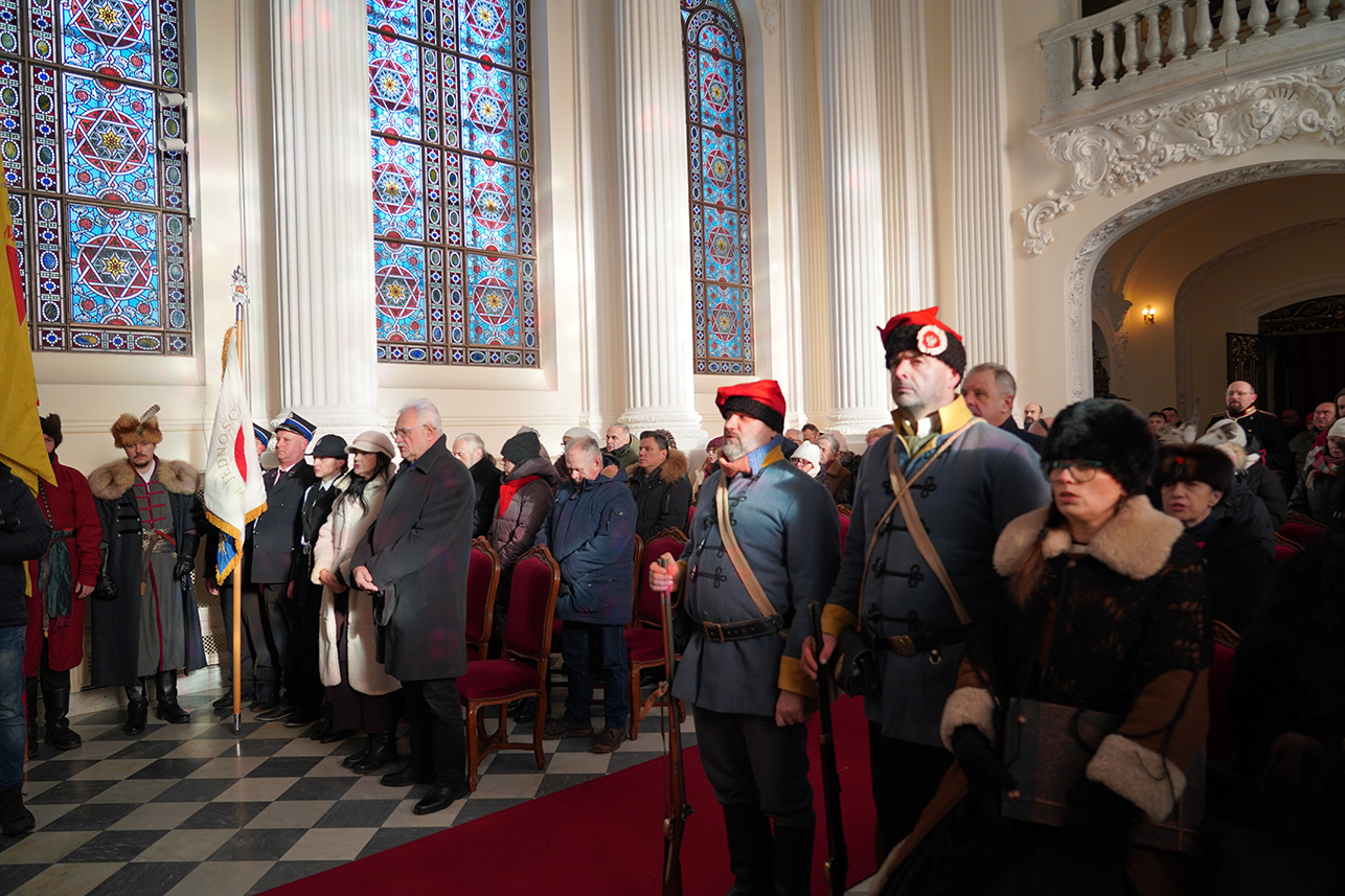 Zdjęcie przedstawia wnętrze wysokiej, reprezentacyjnej sali pałacowej. W tle dominują trzy ogromne okna witrażowe w odcieniach błękitu, czerwieni i bieli, osadzone pomiędzy wysokimi, białymi kolumnami. Witraże rzucają na ściany delikatne, kolorowe refleksy. Podłoga jest czarno-biała, ułożona w szachownicę. Po prawej stronie widoczny jest fragment dekoracyjnego balkonu i arkady prowadzącej do kolejnego pomieszczenia, gdzie palą się ścienne lampy.
W sali stoi liczna grupa osób. Większość to dorośli i seniorzy ubrani w zimowe płaszcze, czapki i szaliki. Uczestnicy zgromadzenia stoją w rzędach, zwróceni w jednym kierunku, jak podczas uroczystości religijnej lub oficjalnej.
Na pierwszym planie, po prawej stronie, widać dwie postacie w historycznych mundurach. To mężczyźni ubrani w szaroniebieskie, długie kaftany przewiązane pasami i skórzanymi rapciami. Na głowach mają wysokie czapki z czarnego futra, jedna z nich jest ozdobiona czerwonym elementem. Jeden z mężczyzn trzyma broń długą przy nodze, drugi stoi wyprostowany, z poważnym wyrazem twarzy.
Obok nich, bliżej prawego brzegu kadru, stoi kobieta w ciemnym płaszczu z jasnym futrzanym kołnierzem, w futrzanej czapce i okularach. W rękach trzyma torebkę.
Po lewej stronie fotografii widoczni są kolejni rekonstruktorzy w strojach historycznych: mężczyźni w długich płaszczach, z futrzanymi czapami i elementami umundurowania. Wśród nich stoi osoba trzymająca biały sztandar na drzewcu. W środkowej części sali ustawiono rzędy czerwonych krzeseł, częściowo zajęte lub odsunięte, a część osób stoi między nimi.
Całość kadru oddaje atmosferę podniosłej uroczystości historyczno-patriotycznej odbywającej się w pałacowym wnętrzu.
