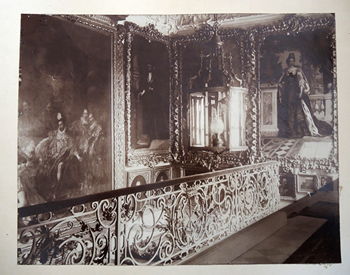 Archiwalne zdjęcie w sepii, ukazujące piętro reprezentacyjnej klatki schodowej pałacu w Kozłówce, około 1900 roku. Wzdłuż posadzki zamocowana jest balustrada dekorowana liśćmi akantu i pośrodku herbem rodziny Zamoyskich, Jelita, przedstawiającym trzy skrzyżowane kopie, zwieńczonym dziewięciopałkową koroną hrabiowską. Poręcz wyłożona materiałem. Nad balustradą widoczna duża wisząca latarnia o metalowej konstrukcji i kryształowych taflach. Wewnątrz jasny szklany klosz. W tle na ścianach obrazy oprawione w dekorowane, złocone ramy.