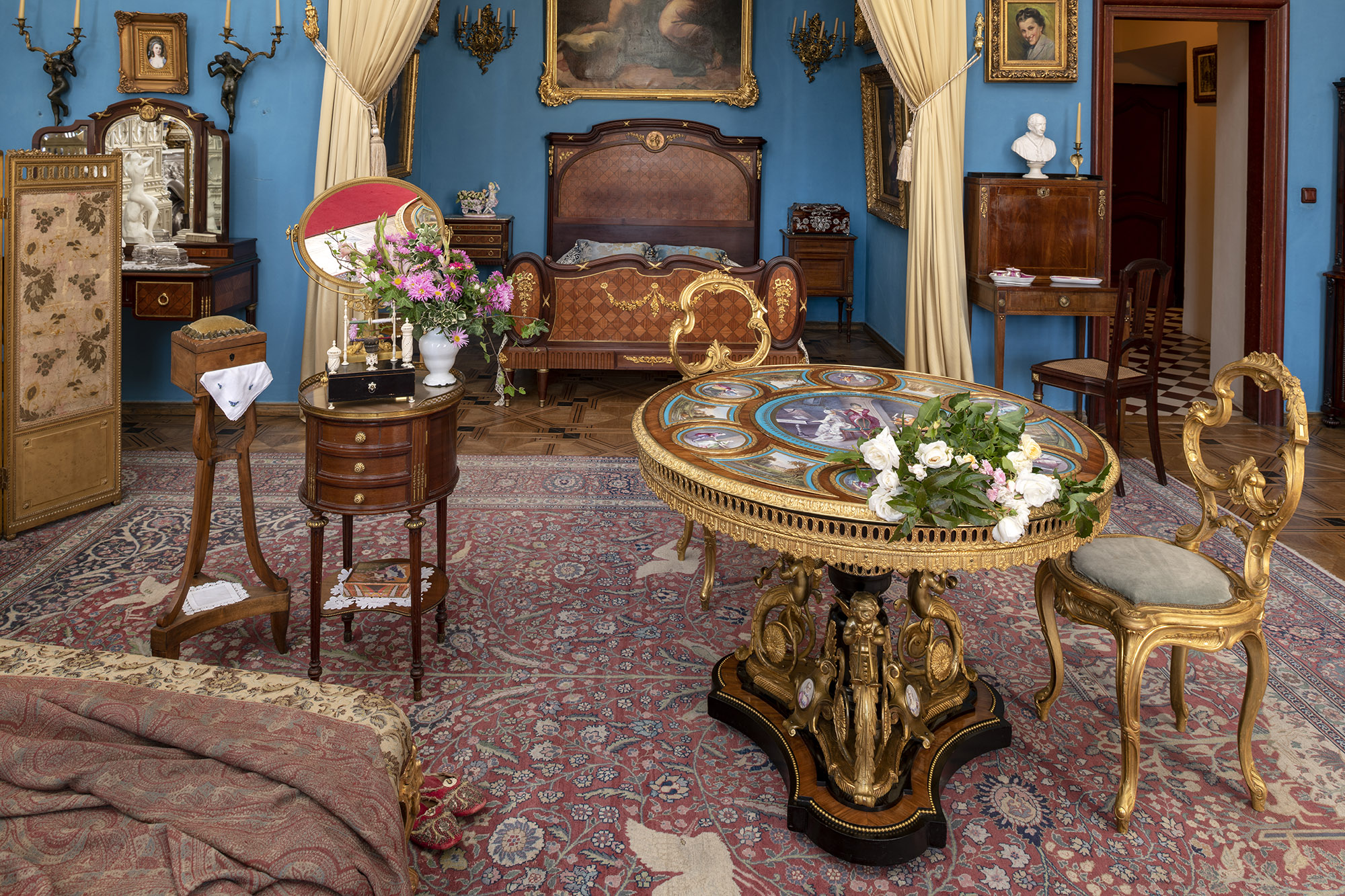 Współczesna kolorowa fotografia przedstawiająca wnętrze Sypialni Hrabiny, nazywanej od koloru ścian Salonem Błękitnym. W głębi widoczne jest ustawione we wnęce podwójne łoże dekorowane intarsją, nad łożem fragment obrazu oprawionego w złoconą ramę. Wejście do wnęki udekorowane jest kotarami, z jasnego połyskliwego materiału spiętego ozdobnymi sznurami zwieńczonymi dużymi chwostami. Po prawej stronie niewielkie wysokie damskie biurko, na nim białe popiersie papieża, powyżej portret z 1941 roku przedstawiający uśmiechniętą Jadwigę Zamoyską, ostatnią właścicielkę Kozłówki. Dalej w prawo otwarte drzwi, widoczna biało czarna mozaika na posadzce i kolejne drzwi. Po lewej stronie wnęki sypialnianej widoczny jest fragment toaletki z lustrem, z ciemnego mahoniu, nad nią kinkiet świecowy z pełnoplastyczną postacią kobiety. Na pierwszym planie fragment szezlongu, przy nim stolik i niciak. Na blacie stolika bukiet różowych kwiatów w jasnym wazonie i lustro, na półeczce poniżej szuflad kolorowe pudełko po słodyczach ułożone na białej serwetce. Po prawej stronie stół udekorowany złoconym brązem i porcelanowymi plakietami.