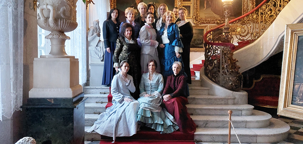 : Kolorowa fotografia przedstawiająca grupę kobiet ubranych w ozdobne suknie. Grupa stoi na schodach. W tle reprezentacyjna klatka schodowa w pałacu. Na ścianach obrazy w złoconych ramach.