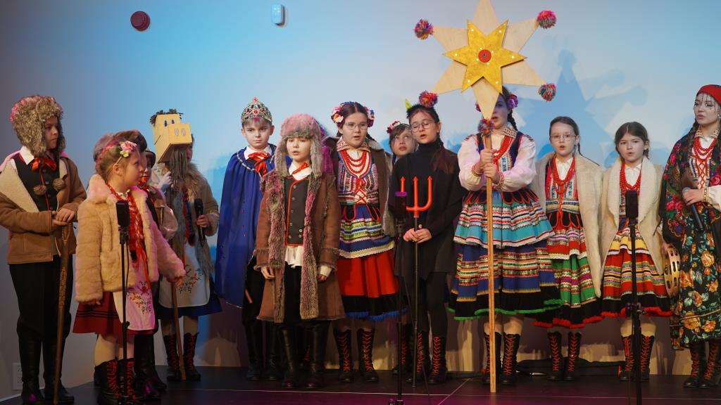 Zespół Pieśni u tańca „Sokół” na scenie w Muzeum Zamoyskich w Kozłówce. Na zdjęciu widoczne są dzieci ubrane w urokliwe ludowe stroje, które bogato odzwierciedlają lokalną tradycję i kulturę. Dzieci noszą barwne hafty, ozdobne naszywki oraz charakterystyczne dla danego regionu elementy folklorystyczne. Niektóre z nich trzymają w rękach symboliczne przedmioty związane z obrzędem kolędniczym.
