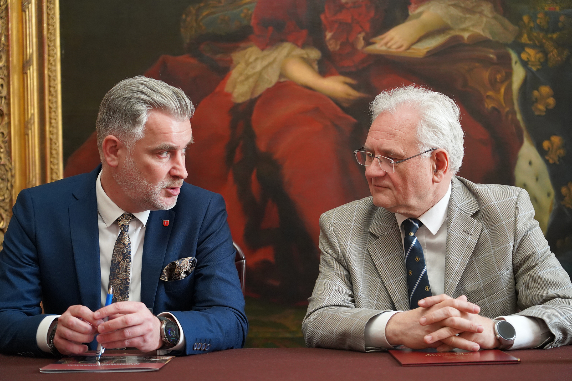 Podpisanie porozumienia współpracy między Muzeum Zamoyskich w Kozłówce a Gminą Kamionka (na zdjęciu od lewej: Pan Karol Ługowski – Burmistrz Miasta Kamionka, Pan Andrzej Pruszkowski – p.o. Dyrektora Muzeum) Na zdjęciu dwóch mężczyzn siedzi obok siebie przy stole i prowadzi rozmowę. Obaj są elegancko ubrani – mężczyzna po lewej w granatowym garniturze, a po prawej w jasnej marynarce w kratę. Ich spojrzenia są skupione i zaangażowane, co sugeruje poważną, ale konstruktywną rozmowę. W tle znajduje się duży, barokowy obraz przedstawiający kobietę w czerwonej sukni, co nadaje całej scenie podniosłego charakteru.