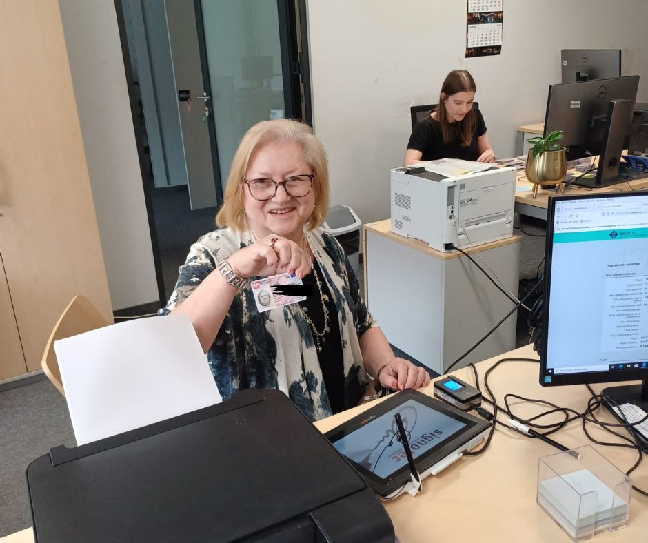 Na pierwszym planie widoczna jest uśmiechnięta starsza kobieta z jasnymi blond włosami i okularami, siedząca przy biurku w nowoczesnym urzędzie. Ma na sobie czarno-białą bluzkę z niebieskimi akcentami i srebrną biżuterię. Kobieta trzyma w dłoni nowo otrzymany polski dowód osobisty i z dumą pokazuje go do kamery. Jej twarz wyraża radość i wzruszenie.
Na biurku przed nią znajduje się monitor komputera, tablet do składania podpisu elektronicznego, drukarka i inne urządzenia biurowe. W tle widać kolejne stanowiska z komputerami i młodą kobietę w czarnej koszulce, pracującą przy jednym z nich. Pomieszczenie jest jasne, nowoczesne, urządzone w stonowanych kolorach.
Zdjęcie dokumentuje ważny moment – odbiór dowodu osobistego.