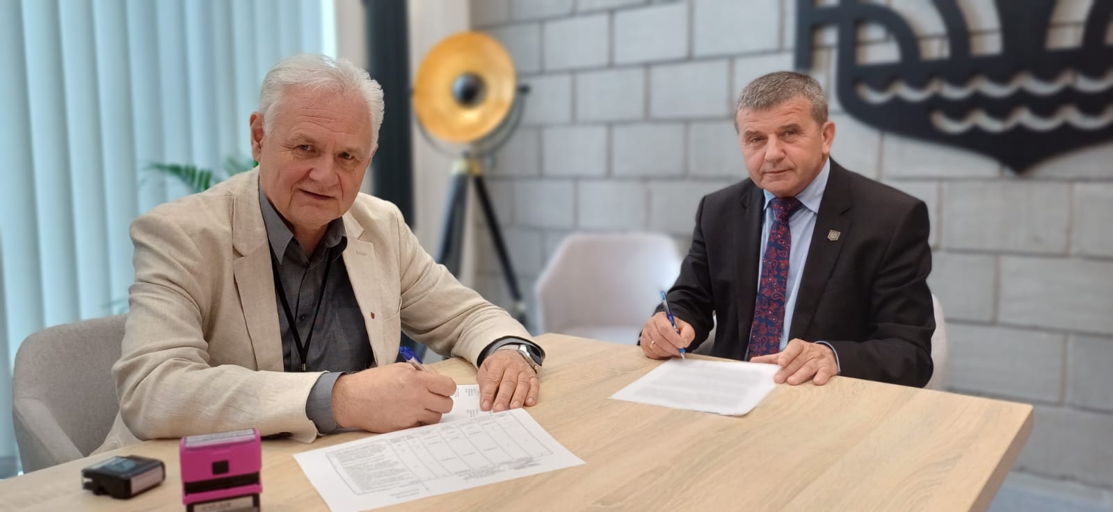 Podpisanie umowy Na zdjęciu widzimy dwóch mężczyzn siedzących przy jasnym, drewnianym stole, na którym leżą dokumenty do podpisania.
• Po lewej stronie siedzi starszy mężczyzna o siwych włosach, ubrany w jasną marynarkę i ciemnoszarą koszulę. Spogląda lekko w stronę fotografującego, trzyma w prawej dłoni długopis, którym podpisał dokument. Obok leżą dwa stempelki – jeden różowy, drugi czarny.
• Po prawej stronie przy stole siedzi drugi mężczyzna w ciemnym garniturze i koszuli oraz we wzorzystym krawacie. Również trzyma długopis i właśnie składa podpis na arkuszu papieru.
W tle za nimi widać ścianę z jasnych, prostokątnych cegieł oraz fragment dużego, czarnego logo umieszczonego na ścianie – ornamentu, który może symbolizować instytucję lub urząd. Po lewej za pierwszym mężczyzną stoi wysoka, trójnogowa lampa z okrągłym, złotym kloszem. Po prawej widać rozmyty zarys jasnego fotela. Cała scena jest dobrze oświetlona – naturalne światło pada zza lewej strony (kierunek zasłonięty przez delikatne, pionowe żaluzje). Atmosfera zdjęcia jest formalna i rzeczowa, sugerująca uroczysty moment podpisywania ważnego porozumienia lub umowy.