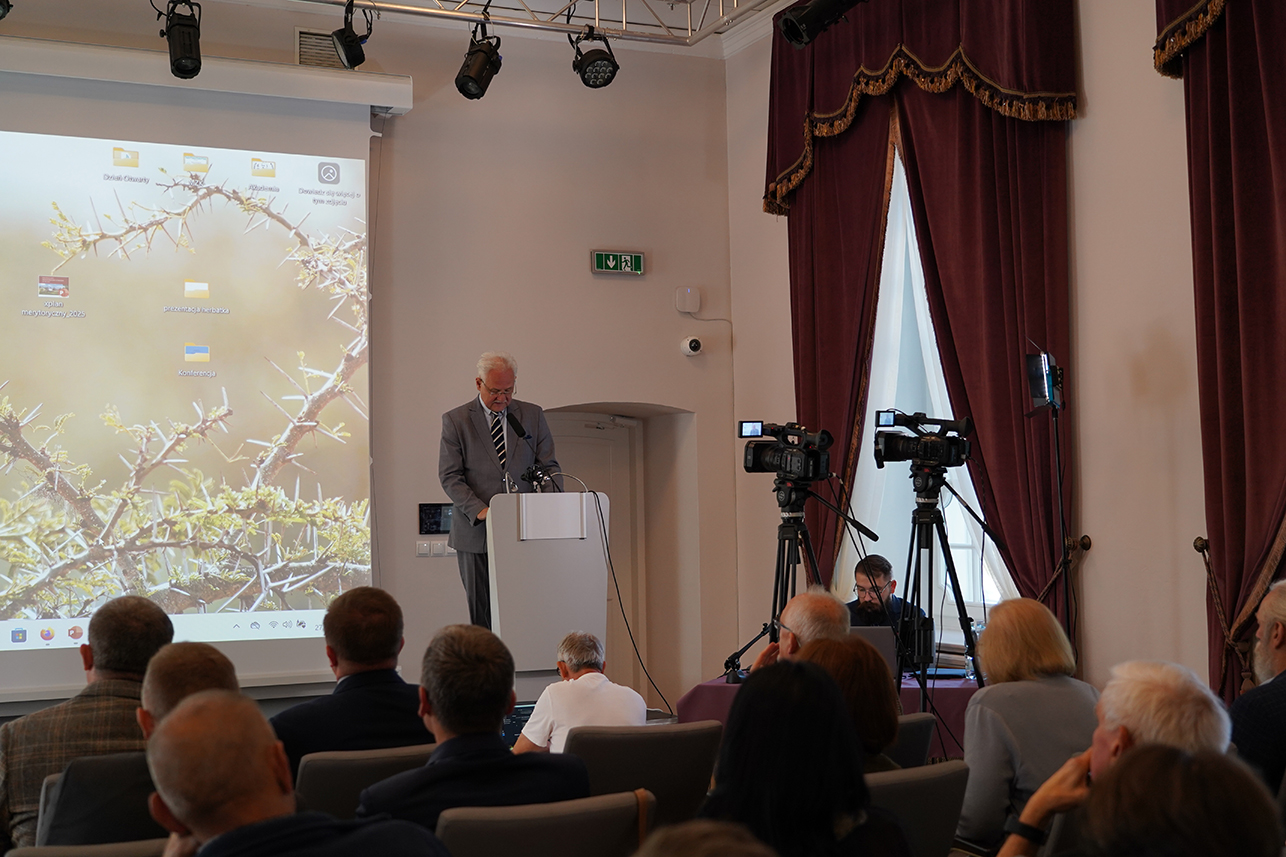 Konferencja naukowa W eleganckiej sali konferencyjnej trwa wystąpienie. Na podwyższeniu za białą mównicą stoi mężczyzna w szarym garniturze i krawacie w paski, jest to p. Andrzej Pruszkowski, Dyrektor Muzeum Zamoyskich w Kozłówce. Pochyla się nad notatkami lub mikrofonem. Za nim rozciąga się wielki ekran z rzutnikiem – widać na nim pulpit komputera z kilkoma ikonami folderów.
Po prawej stronie ustawiono dwie kamery na statywach, skierowane na mównicę. Obok nich siedzi technik przy stole, obsługujący sprzęt. Sala jest pełna – na pierwszym planie widać plecy publiczności, kilkanaście osób siedzi w rzędach na krzesłach. Ściany zdobią wysokie okna z ciężkimi, bordowymi zasłonami obszytymi złotą lamówką. Atmosfera jest oficjalna i poważna – przypomina konferencję prasową lub spotkanie naukowe.