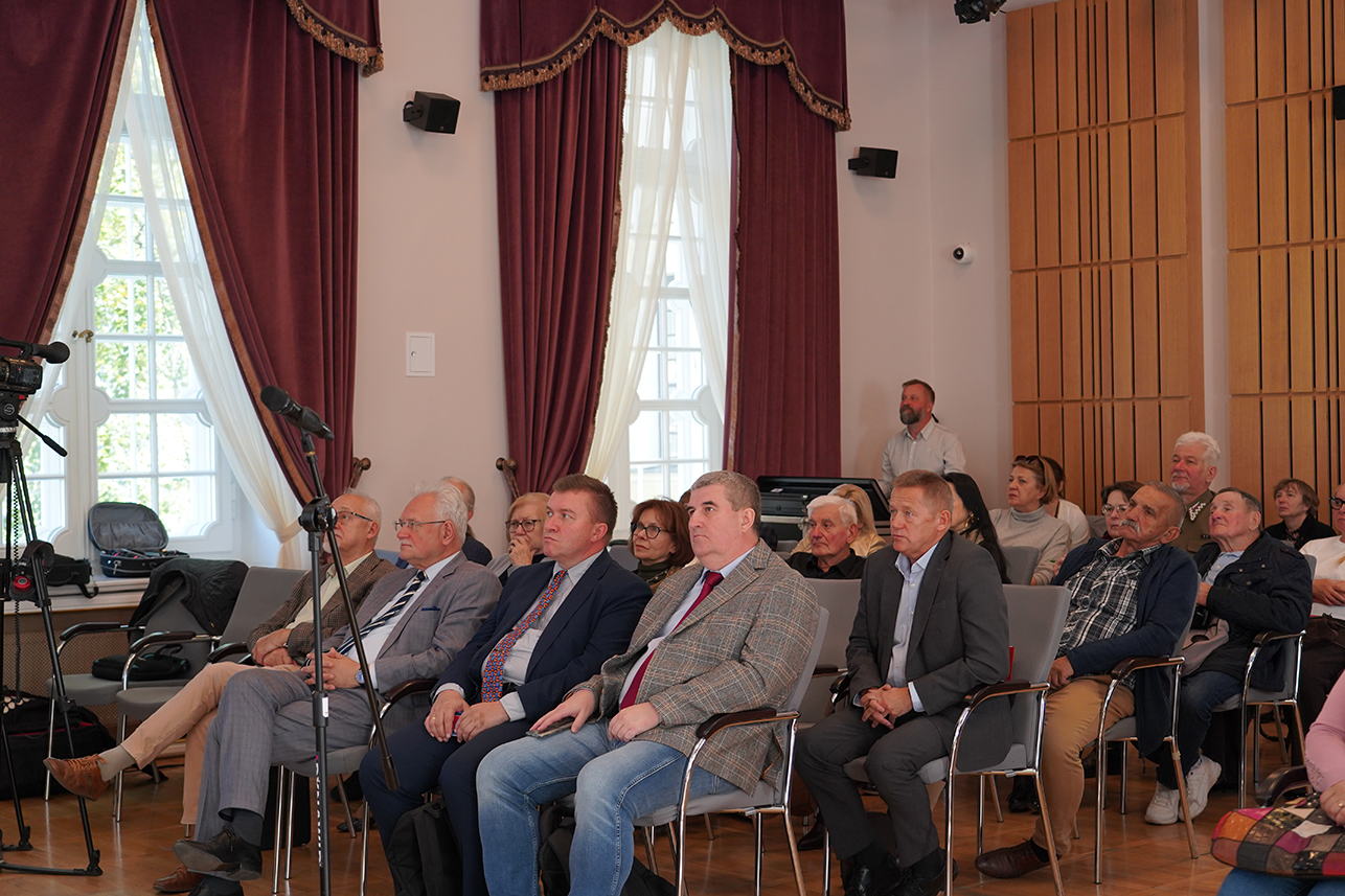 Konferencja naukowa Zdjęcie przedstawia grupę osób siedzących w sali konferencyjnej. Na pierwszym planie, w rzędzie szarych krzeseł, widzimy pięciu mężczyzn w garniturach i marynarkach. Dwaj z nich mają na sobie ciemne garnitury z krawatami, a pozostali ubrani są w marynarki w kratę – jeden z nich zestawił ją z jasną koszulą i dżinsami. Wszyscy siedzą spokojnie, patrząc w stronę wydarzenia poza kadrem.
Za nimi widoczne są kolejne osoby – zarówno mężczyźni, jak i kobiety – ubrane elegancko, w stylu formalnym. Po lewej stronie kadru, w tle, stoją bordowe, ciężkie zasłony i jasne okno, przez które wpada światło dzienne. W prawym górnym rogu, przy stole technicznym, mężczyzna obsługuje sprzęt. Atmosfera jest skupiona, a publiczność wygląda na uważnie wsłuchaną w przebieg spotkania.
