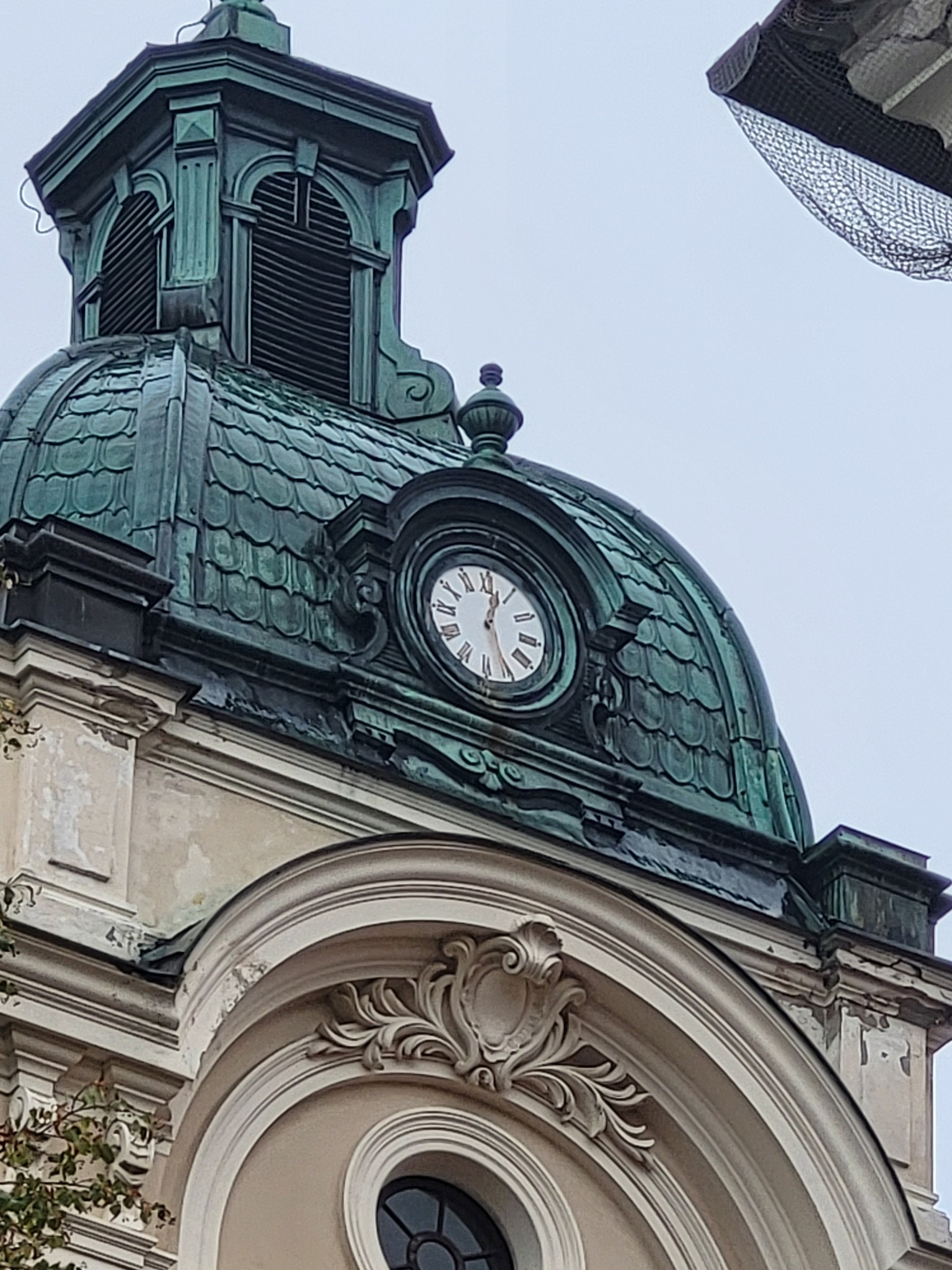 Na zdjęciu widoczny jest fragment zabytkowego budynku, ujęty z dołu, tak jakby fotograf stał na ulicy i patrzył w górę. Kadr wypełnia przede wszystkim kopuła dachu pokryta metalową blachą o zielonkawym, spatynowanym kolorze. Powierzchnia kopuły ma dekoracyjny wzór przypominający łuski lub dachówki, a na jej łączeniach widać pionowe pasy wzmacniające konstrukcję.
Centralnie, na przedniej części kopuły, znajduje się okrągły zegar w bogato zdobionej oprawie. Tarcza jest jasna, z rzymskimi cyframi, a wskazówki ustawione są na godzinę około dwunastej (zbliżone do 12:00). Zegar otacza masywna, ciemniejsza rama z ornamentami, a nad nim wystaje niewielka ozdobna sterczyna zakończona kulką.
Ponad kopułą wznosi się niewielka wieżyczka z otwartymi arkadami i czarnymi żaluzjowymi przesłonami, sugerująca dzwonnicę lub element wentylacyjny. Wieżyczka ma ten sam zielony odcień patyny i geometryczne zdobienia na narożach. U góry jest zwieńczona małym daszkiem i elementem dekoracyjnym.
Niżej, pod kopułą, widać jasną, kremowo-beżową elewację z miejscami odpryskującym tynkiem. Nad dużym łukowym oknem znajduje się dekoracyjny kartusz i rokokowe ornamenty z liściastymi motywami. Samo okno jest częściowo widoczne – ciemne, z podziałami.
Tło stanowi jednolite, jasnoszare niebo. W prawym górnym rogu kadru pojawia się fragment siatki lub osłony oraz kawałek innej konstrukcji, a w lewym dolnym rogu widać niewielkie gałązki z liśćmi, które delikatnie wchodzą w kadr. Całość ma chłodny, stonowany klimat, podkreślony przez patynowaną zieleń metalu i pochmurne światło.