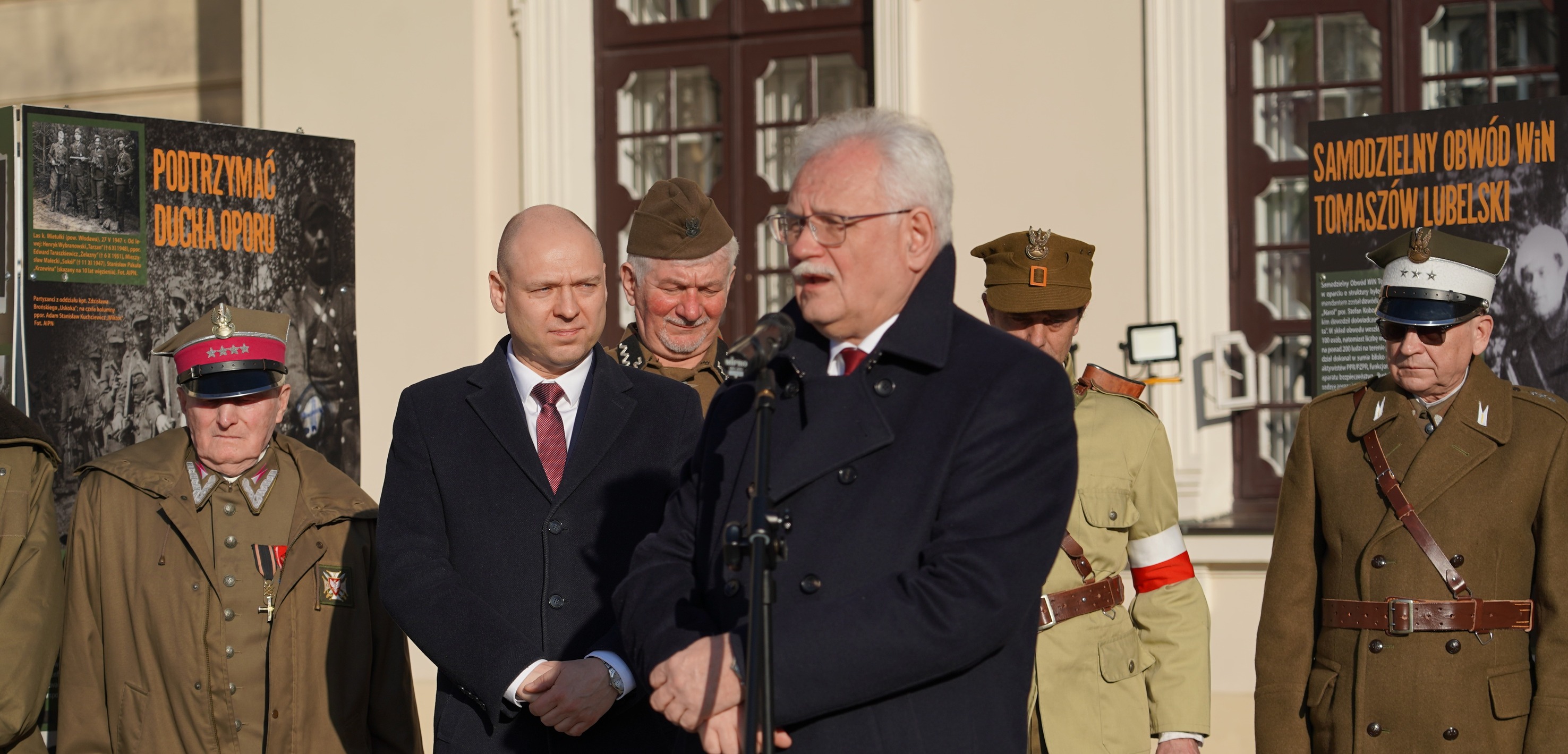 Na fotografii widoczna jest uroczystość plenerowa przed jasnym, reprezentacyjnym budynkiem z dużymi, ciemnobrązowymi oknami i białymi obramowaniami. Jest słonecznie; światło pada z prawej strony kadru, tworząc wyraźne cienie na twarzach i ubraniach.
Na pierwszym planie, nieco po prawej, stoi mężczyzna w średnim lub starszym wieku, o siwych włosach, w okularach, w ciemnym, dwurzędowym płaszczu. Przemawia do mikrofonu ustawionego na statywie; ręce ma złożone przed sobą. Jego twarz jest skupiona, jak w trakcie oficjalnego wystąpienia.
Za nim, po lewej stronie, stoi łysy mężczyzna w ciemnym płaszczu lub garniturze, z białą koszulą i bordowym krawatem. Ma poważny wyraz twarzy i patrzy w kierunku mówcy. Między nimi, w głębi, widać uśmiechniętego starszego mężczyznę w brązowej, historycznej czapce i mundurowej stylizacji.
Po bokach kadru stoją mężczyźni w rekonstrukcyjnych lub historycznych mundurach. Po lewej, na skraju zdjęcia, znajduje się starszy mężczyzna w oliwkowym płaszczu mundurowym i czapce z jasnym otokiem i czerwonym pasem; na piersi ma odznaczenia. Po prawej stronie kadru stoi kolejny mężczyzna w brązowym mundurze z szerokim, skórzanym pasem przewieszonym przez pierś; ma jasną czapkę z daszkiem i ciemne okulary. Za mówcą, po prawej, częściowo widoczny jest jeszcze jeden uczestnik w jasnym mundurze z czerwonobiało-czerwoną opaską na rękawie.
W tle, po obu stronach grupy, ustawiono dwa duże planszowe plakaty na stojakach. Na lewym plakacie, na tle czarno-białych zdjęć, widnieje duży pomarańczowy napis: „PODTRZYMAĆ DUCHA OPORU”. Na prawym plakacie, również z archiwalnymi fotografiami, znajduje się pomarańczowy tytuł: „SAMODZIELNY OBWÓD WiN TOMASZÓW LUBELSKI”. Całość ma charakter oficjalnej ceremonii historyczno-patriotycznej: przemówienie, obecność rekonstruktorów i plansz informacyjnych.