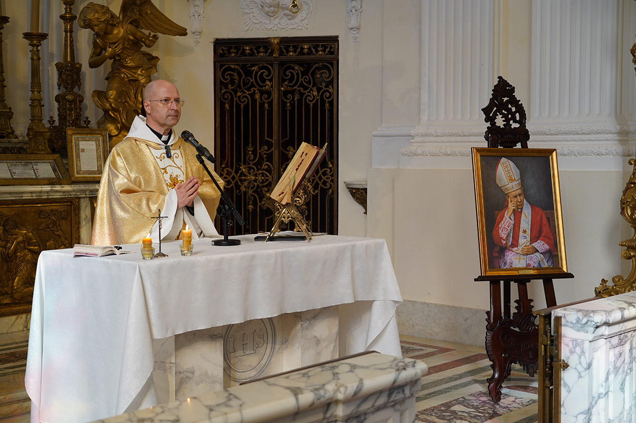 We wnętrzu kaplicy przy marmurowym ołtarzu stoi ksiądz w złotej szacie liturgicznej. Przed nim znajduje się mikrofon i otwarta księga na pulpicie, a na ołtarzu palą się świece. Obok ołtarza, na drewnianym stojaku, ustawiony jest obraz przedstawiający kardynała w czerwonych szatach. W tle widoczne są jasne kolumny oraz elementy bogato zdobionego wystroju kaplicy.