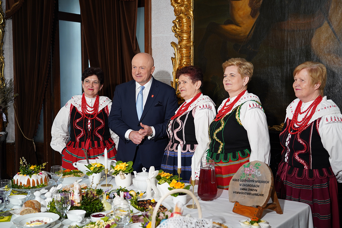 W eleganckim wnętrzu przy nakrytym stole stoi grupa osób pozujących do zdjęcia. Pośrodku znajduje się mężczyzna w garniturze, a obok niego kilka kobiet ubranych w tradycyjne stroje ludowe z czerwonymi koralami.
Na stole ustawione są różnorodne potrawy, naczynia i dekoracje, w tym kwiaty oraz dzbanek z napojem. W tle widoczny jest duży obraz w ozdobnej ramie oraz ciężkie zasłony. Całość ma uroczysty i regionalny charakter.