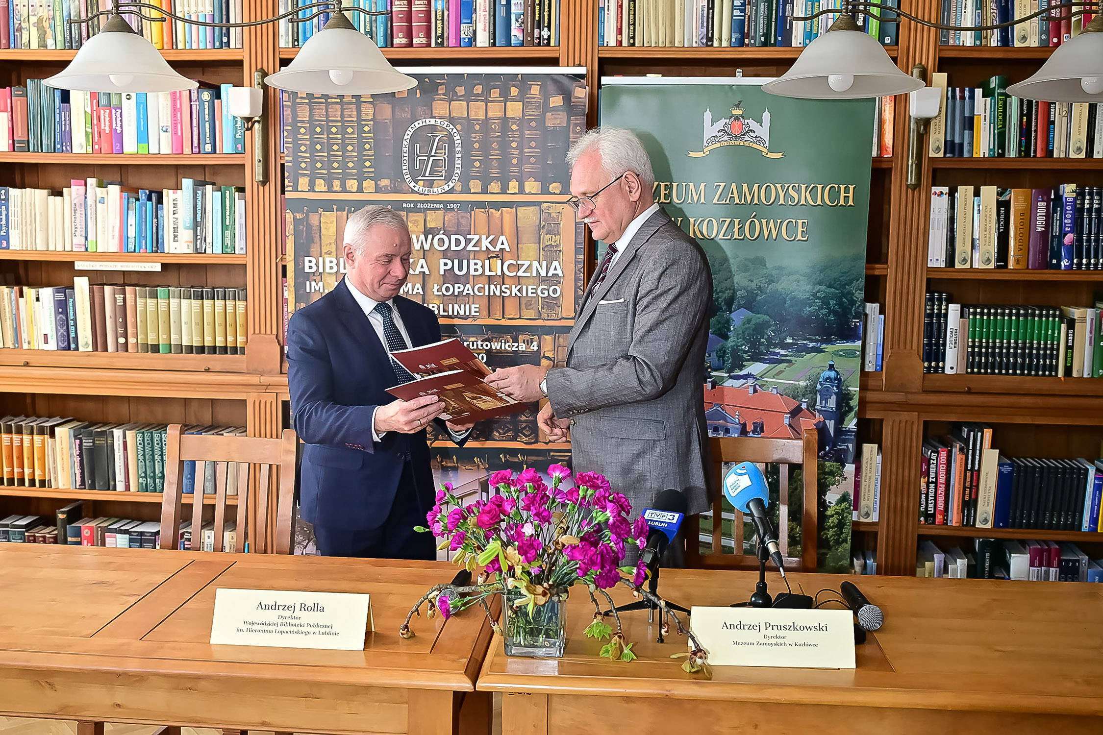 Na zdjęciu widoczna jest scena z konferencji prasowej odbywającej się w czytelni biblioteki. W centralnej części kadru stoją naprzeciw siebie dwaj mężczyźni w eleganckich garniturach – dyrektorzy instytucji podpisujących umowę o współpracy. Jeden z nich trzyma otwarty dokument w czerwonej oprawie, który wspólnie przeglądają.
Za nimi znajdują się wysokie, drewniane regały wypełnione książkami. Po obu stronach ustawiono roll-upy z nazwami i logotypami instytucji: Wojewódzkiej Biblioteki Publicznej w Lublinie oraz Muzeum Zamoyskich w Kozłówce.
Na pierwszym planie stoi drewniany stół konferencyjny. Ustawiono na nim mikrofony oraz wizytówki z imionami i nazwiskami dyrektorów. Pośrodku stołu znajduje się niewielki bukiet różowych kwiatów w szklanym wazonie.